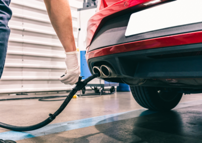 A mechanic in gloves prepares to service the exhaust of a red car in a workshop, focusing on maintenance tasks.