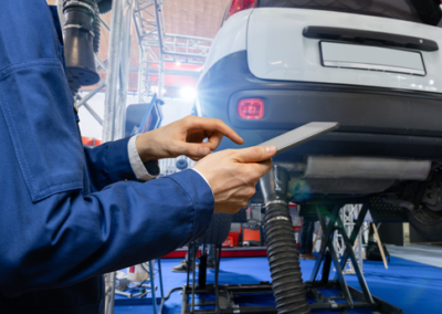 A mechanic in a blue uniform uses a tablet to diagnose a car raised on a lift in a well-lit automotive workshop.