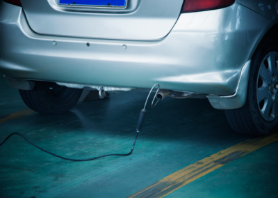 A silver car's rear with an attached probe for emissions testing, resting on a green garage floor with yellow lines.