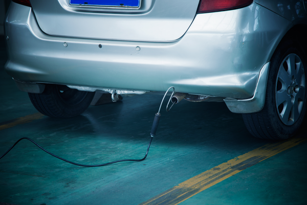 A silver car's rear with an attached probe for emissions testing, resting on a green garage floor with yellow lines.