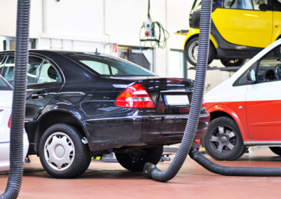 A black sedan in an auto repair shop is surrounded by various cars, with exhaust hoses positioned nearby.