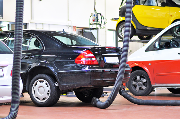 94915373_l A black sedan in an auto repair shop is surrounded by various cars, with exhaust hoses positioned nearby.